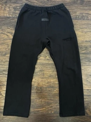 Svarta mjukisbyxor från Essentials - Svarta mjukisbyxor från Essentials med diskret logga på vänster lår och elastisk midja. Byxorna har raka ben och sidofickor, perfekta för en avslappnad och trendig stil. Tillverkade i mjukt material för maximal komfort.