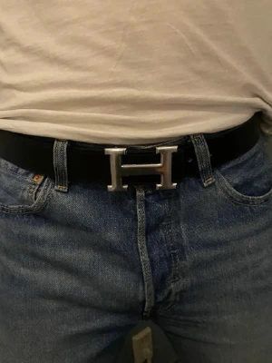 H vändbart skärp med H-spänne - Snyggt vändbart skärp från H med ikoniskt H-spänne i silver. Ena sidan är svart och andra sidan är vit, vilket gör det enkelt att matcha med olika outfits. Skärpet är gjort i exklusivt skinn och har en stilren, klassisk design.