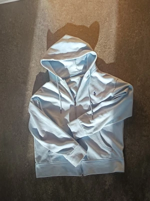 Ljusblå hoodie Polo Ralph Lauren - Snygg ljusblå hoodie med dragkedja Polo Ralph Lauren. Klassisk broderad logga på bröstet, justerbar huva med snören och två fickor framtill. Perfekt för en chill och stilren look.