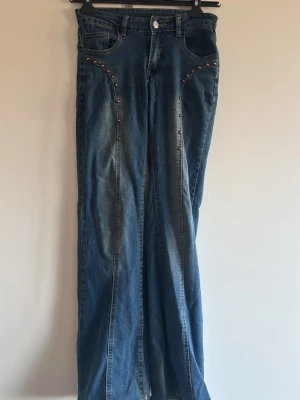 Blå utsvängda jeans med nitar - Säljer ett par blå jeans med utsvängda ben och coola nitar längs fickorna framtill. Jeansen har klassisk femficksmodell och en snygg tvättad look. Perfekta för dig som vill sticka ut lite extra med detaljer. Materialet är denim i bomull.
