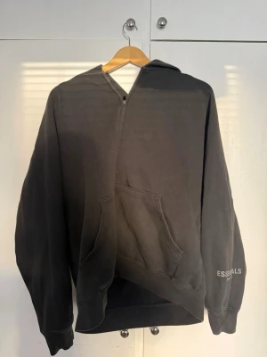 Svart Essentials Fear of God hoodie - Säljer en svart hoodie från Fear of God Essentials med halv dragkedja och stor känguruficka framtill. Trycket 'ESSENTIALS FEAR OF GOD' finns på ärmen. Hoodien har en relaxed fit och är tillverkad i mjukt bomullsmaterial. Perfekt för en chill och stilren look.