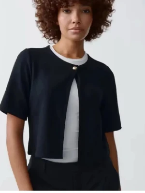 Svart kortärmad kofta med knapp - Stilren svart kofta med korta ärmar och en dekorativ pärlknapp framtill. Koftan är croppad och har rund halsringning. Perfekt att slänga över en t-shirt för en trendig look.