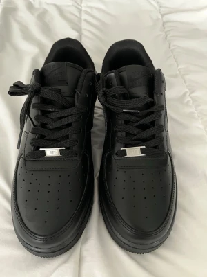 Nike Air Force 1 helsvarta sneakers - Snygga helsvarta Nike Air Force 1 sneakers i klassisk low-modell. Skorna är tillverkade i läder med matchande svarta skosnören och diskret Nike-logga på sidan. Perfekta för dig som gillar en clean och stilren look.