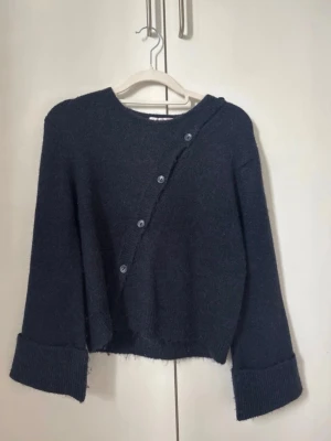 Mörkblå stickad tröja med knappar - Mysig mörkblå stickad tröja med sned knäppning framtill och stora uppvikta ärmslut. Tröjan har en boxig passform och är tillverkad i ett mjukt, luddigt material. Perfekt för lager-på-lager och chill dagar. Den är helt ny utan prislapp