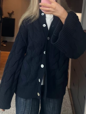 Mörkblå kabelstickad kofta Gina Tricot  - Säljer en mörkblå kabelstickad kofta med stora vita knappar. Koftan har rund halsringning, breda ribbade muddar och ett klassiskt flätmönster. Perfekt för lager-på-lager och riktigt mysig att ha på sig.