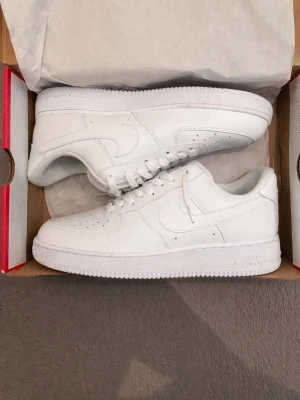 Nike Air Force 1 vita sneakers - Klassiska helvita Nike Air Force 1 sneakers i läder med perforerad tå, vit sula och ikonisk swoosh på sidorna. Skorna har lågt skaft och plös med Nike-logga. Perfekta för en clean och stilren look.