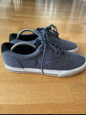 Grå sneakers från Polo Ralph Lauren - Stilrena grå sneakers från Polo Ralph Lauren med ovandel i filtliknande tyg och svarta detaljer vid häl och plös. Skorna har vita sulor med diskret rand och klassisk snörning. Perfekta för en avslappnad och trendig look.Sparsamt använda.  Storlek 43 herrskor. Har alltid haft skoblock i så den har hållit formen väl.