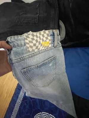 Ljusblå ripped jeans från FSBN - Säljer ett par ljusblå jeans från FSBN med slitna detaljer och snygg tvätt. Jeansen har klassisk femficksdesign och en schackrutig patch bak i midjan. Perfekta för en avslappnad och trendig look.