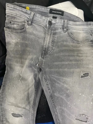Grå slitna jeans från Smog Denim - Snygga grå jeans från Smog Denim med slitningar och hål på benen samt vita färgstänk för en trendig look. Jeansen har klassisk femficksmodell och raka ben. Perfekta för dig som gillar streetstyle och en avslappnad vibe.