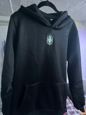Svart hoodie med Brasilien-emblem - Svart hoodie med Brasilien-emblem på bröstet och klassisk huva. Tröjan har en stor magficka och ribbade muddar. Perfekt för dig som gillar sportiga och stilrena plagg. Materialet känns mjukt och bekvämt, passar till chill eller träning.