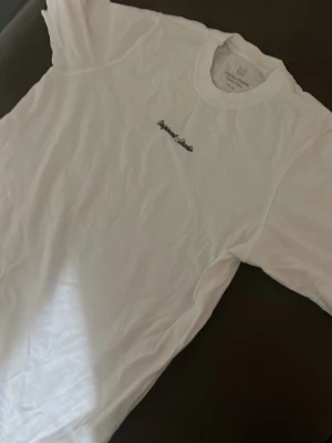 Vit t-shirt från Jack & Jones - Säljer en klassisk vit t-shirt från Jack & Jones med diskret broderad text på bröstet. T-shirten har rund hals och korta ärmar, perfekt för en clean och enkel stil. Tillverkad i mjuk bomull för skön komfort.