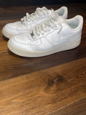 Vita Nike Air Force 1 sneakers - Klassiska vita Nike Air Force 1 sneakers i läder med låg profil och perforerad tå för ventilation. Swoosh-logga på sidan och präglad 'AIR'-text på sulan. Tjock vit yttersula och matchande vita skosnören. Perfekt för en clean och tidlös streetwear-look.