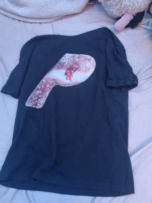 Svart Palace t-shirt med grafiskt tryck - Svart t-shirt från Palace med ett stort grafiskt tryck på framsidan som föreställer ett P-format motiv med rosa och vita detaljer. Klassisk passform och rund hals. Perfekt för dig som gillar streetwear och unika prints.