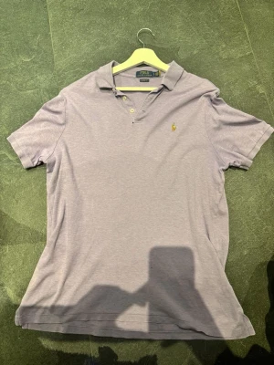Ljuslila pikétröja från Polo Ralph Lauren - Klassisk ljuslila pikétröja från Polo Ralph Lauren med korta ärmar och krage. Tröjan har knappar vid halsen och den ikoniska broderade loggan på bröstet. Perfekt till jeans eller shorts för en clean och stilren look.
