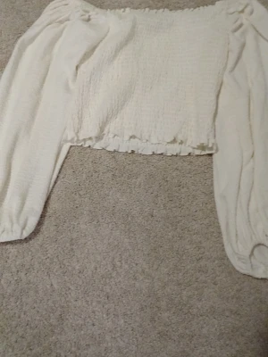 Vit offshoulder topp från Iconic - Trendig vit offshoulder topp från Iconic med långa puffärmar och smockad, croppad passform. Toppen har volangkant nedtill och är perfekt för dig som gillar en romantisk och luftig stil. Tillverkad i mjukt material som känns skönt mot huden.