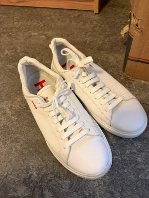 Vita sneakers från Hugo - Stilrena vita sneakers från Hugo med klassisk låg profil och vita snören. Skorna har en diskret röd Hugo-logga på sidan och stor logotyp på innersulan. Tillverkade i slätt läder med vit sula och enkel design som passar till det mesta.