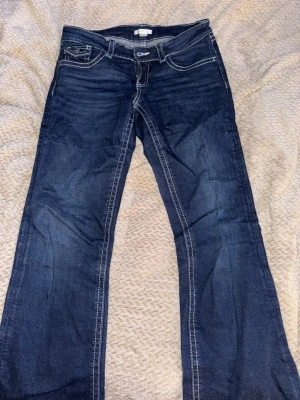 Mörkblå bootcut jeans med kontrastsömmar - Snygga mörkblå bootcut jeans med vita kontrastsömmar och klassiska fem fickor. Jeansen har en knappstängning och detaljerad bakficka med lock. Perfekta för dig som gillar en tidlös och avslappnad stil.
