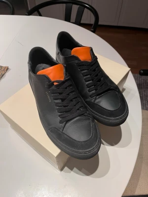 Svarta Axel Arigato sneakers med orange detalj - Snygga svarta sneakers från Axel Arigato med en cool orange detalj på plösen. Skorna har svart ovandel i läder, svarta skosnören och en klassisk låg siluett. Perfekta för dig som gillar stilrena men ändå unika sneakers.