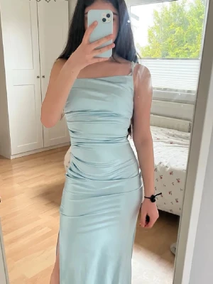 Ljusblå långklänning med slits - Säljer en elegant ljusblå långklänning i glansigt material. Klänningen har smala axelband, figurnära passform och en snygg slits på sidan. Perfekt för dig som vill sticka ut med en stilren look.