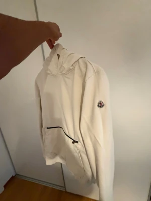 Vit hoodie med Moncler-logga - Snygg vit hoodie från Moncler med klassisk huva och stor magficka med svart dragkedja. Monclers ikoniska logga sitter på vänster ärm. Hoodien har ribbade muddar vid ärmslut och nederkant. Perfekt för en clean och stilren look. (Storlek S passar som M)