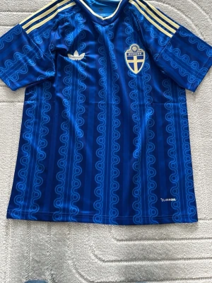 Blå Sverige fotbollströja Adidas - Säljer en blå fotbollströja från Adidas med Sveriges landslagsemblem på bröstet. Tröjan har gula ränder på axlarna, mönstrad design och korta ärmar. Materialet är lätt och andas, perfekt för träning eller match.