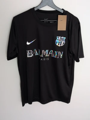 Svart Nike x Balmain t-shirt finns s,m - Snygg svart t-shirt med holografiskt Balmain Paris-tryck, Nike-logga och FCB-emblem på bröstet. Klassisk rund hals och korta ärmar. Perfekt för dig som gillar streetwear och vill sticka ut med en unik design.