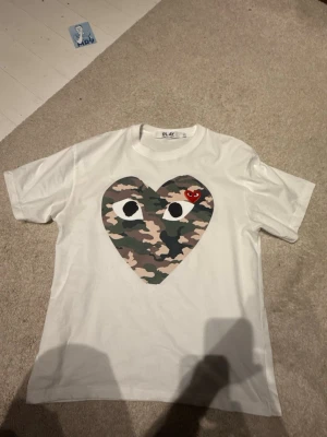 Vit Comme des Garçons PLAY t-shirt - Säljer en vit t-shirt från Comme des Garçons PLAY med stort hjärta i camouflage på bröstet och ett litet rött hjärta med ögon. Klassisk rund hals och korta ärmar. Perfekt statement-plagg för dig som gillar streetwear.