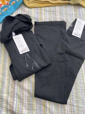 Lululemon Svart träningsset med hoodie och tights - Snyggt svart träningsset med en zip-hoodie och matchande tights. Hoodien har dragkedja framtill och fickor, medan tightsen är tighta och stretchiga. Perfekt för gymmet eller löprundan. Materialet är mjukt och följsamt för maximal komfort.