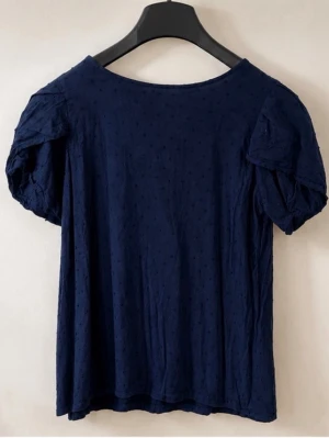 Mörkblå blus  - Söt mörkblå blus med korta puffärmar och diskret prickigt mönster. Blusen har rund halsringning och en knappdetalj bak i nacken. Perfekt att styla med jeans eller kjol för en trendig look.