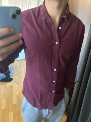 Mörkröd Ralph Lauren skjorta med knappar - Snygg mörkröd skjorta med klassisk krage och vita knappar. Skjortan har lång ärm och en liten broderad logga på bröstet. Perfekt för dig som vill ha en stilren och enkel look.