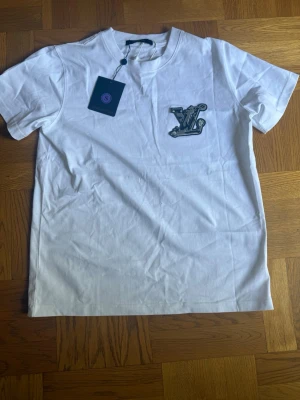 Vit t-shirt med  - Vit t-shirt med kort ärm och rund halsringning. På bröstet finns en broderad i svart och grått. Baksidan har en brun patch upptill. Perfekt för en clean och stilren look.