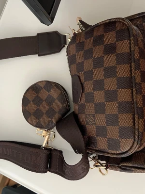 Brun Louis Vuitton axelremsväska - Snygg brun axelremsväska från Louis Vuitton med klassiskt rutmönster i mörkbrunt och ljusbrunt. Väskan har bred axelrem med logga och matchande rund liten coin pouch. Guldiga metalldetaljer och dragkedja. Helt ny. Köpt utomlands 