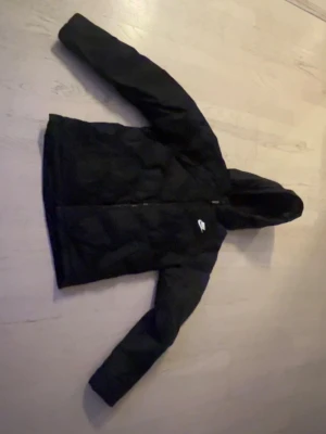 Svart pufferjacka från Nike XL - Säljer en svart pufferjacka från Nike i storlek 158-170. Jackan har huva, dragkedja framtill och en liten Nike-logga på bröstet. Tillverkad i ett slitstarkt material som håller värmen under kalla dagar. Perfekt för vintern och riktigt snygg streetstil.