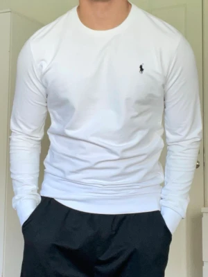 Vit långärmad tröja från Ralph Lauren - Klassisk vit långärmad tröja från Ralph Lauren med rund halsringning och den ikoniska lilla broderade loggan på bröstet. Tröjan är i mjukt material och har en clean, stilren look som passar till det mesta.