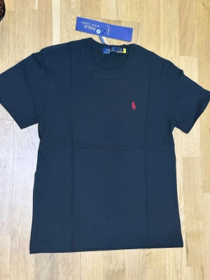 Svart t-shirt från Polo Ralph Lauren - Klassisk svart t-shirt från Polo Ralph Lauren med röd broderad logga på bröstet. T-shirten har rund halsringning och korta ärmar. Tillverkad i mjuk bomull för en skön känsla och enkel stil.
