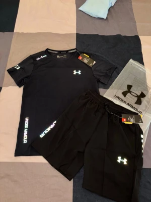 Svart träningsset från Under Armour - Snyggt svart träningsset från Under Armour med t-shirt och shorts. Setet har reflekterande loggor och textdetaljer i regnbågsfärger. Tillverkat i ett lätt och snabbtorkande funktionsmaterial som passar perfekt till gymmet eller löpning.
