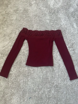 Vinröd offshoulder topp med spets - Trendig vinröd offshoulder topp med långa ärmar och bred spetskant upptill. Toppen är ribbad och har en tight passform som framhäver figuren. Perfekt för dig som vill ha en stilren men ändå romantisk look. Den är insydd, men det är inget som är klippt så det går att sprätta bort.