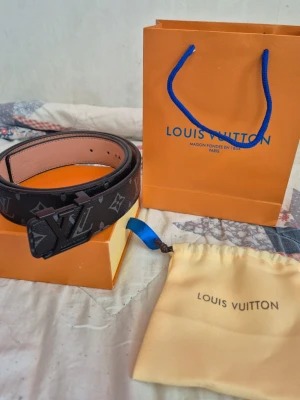 Louis Vuitton bälte  - Snyggt svart bälte från Louis Vuitton med klassiskt monogrammönster och stor LV-logga som spänne. Bältet har en matt finish och detaljer i mörk metall. Perfekt accessoar för att lyfta din outfit.