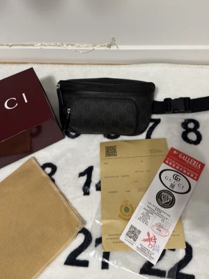 Svart shoppingväska från G - Snygg svart shoppingväska från G med klassiskt mönster i canvas och detaljer i läder. Väskan har justerbart midjeband, dragkedja framtill och flera fack för smart förvaring. Perfekt accessoar för dig som vill ha både stil och funktion.