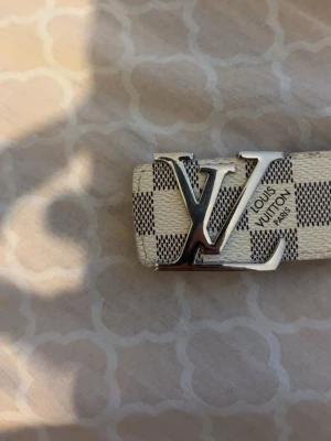  rutigt bälte i beige - Snyggt bälte med klassiskt rutigt mönster i beige och vitt. Bältet har en stor silvrig spänne framtill och är tillverkat i skinn. Perfekt accessoar för att lyfta din outfit.