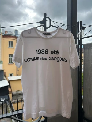 Vit Comme des Garçons t-shirt - Säljer en vit t-shirt från Comme des Garçons med svart tryck '1986 été COMME des GARÇONS' på bröstet. Klassisk rund hals och korta ärmar. Perfekt för dig som gillar stilrena och ikoniska plagg.