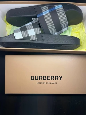 Svarta sandaler med rutigt mönster Burberry - Snygga svarta sandaler från Burberry med klassiskt rutigt mönster i svart, vitt och grått samt en diskret blå linje. Tillverkade i syntetmaterial med bred rem över foten och bekväm sula. Perfekta för en trendig och avslappnad stil.