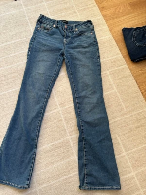 Blå bootcut jeans från True religion med rosa detaljer - Snygga blå bootcut jeans med rosa detaljer. Passformen heter Becca och är Mid Risa bootcut. Nästan helt oanvända. Finns minimalt täckrn på byxorna längst ner. Kan skicka bild vid intresse! 