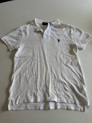 Vit pikétröja från Ralph Lauren - Klassisk vit pikétröja från Ralph Lauren med korta ärmar och krage. Tröjan har den ikoniska lilla loggan broderad på bröstet och knäppning vid halsen. Perfekt till jeans eller shorts för en clean och stilren look. Extremt bra skick då den endast har används ett fåtal gånger. 