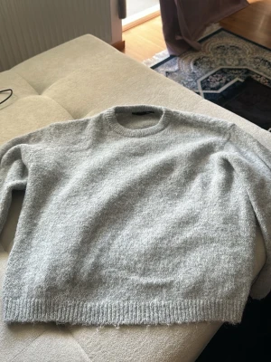 Ljusgrå stickad tröja - Mysig ljusgrå stickad tröja med rund halsringning och ribbade muddar vid ärmslut och nederkant. Tröjan har en enkel och stilren design, perfekt för lager på lager och passar till det mesta. Materialet ser ut att vara mjukt och fluffigt, troligen ull eller en ullmix.