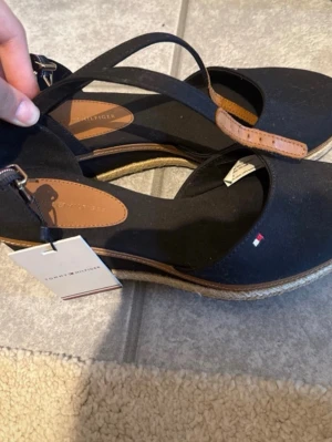 Svarta sandaler från Tommy Hilfiger - Snygga svarta sandaler från Tommy Hilfiger med klassisk espadrille-sula i flätad jute och remmar i textil och läderdetaljer. Liten logga på sidan och justerbart spänne vid ankeln. Perfekta för en trendig och avslappnad look.