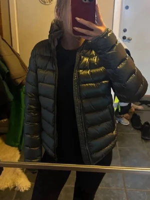Svart dunjacka med huva - Snygg svart dunjacka med huva och quiltad design. Jackan har dragkedja framtill och en patch på ärmen. Perfekt för kalla vinterdagar och håller dig varm med sin fluffiga fyllning. Passar till det mesta och är lätt att matcha med andra plagg.
