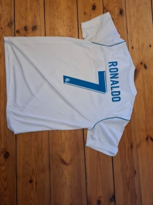 Vit Real Madrid Ronaldo tröja - Säljer en vit fotbollströja med blå detaljer från Real Madrid. På ryggen står det 'RONALDO' och nummer 7 i blått. Tröjan har korta ärmar och är tillverkad i ett lätt, andningsbart material som passar perfekt till träning eller match.
