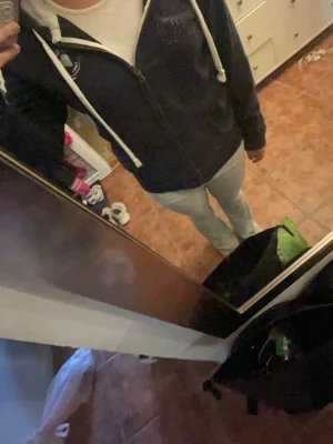 Mörkblå hoodie med dragkedja - Mörkblå hoodie med vit dragkedja och vita snören i huvan. Har två fickor framtill och ett tryckt emblem på bröstet samt dekorativa stenar på andra sidan. Mjuk insida och lång ärm, perfekt för chill dagar.