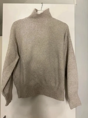 Beige stickad polotröja - Mysig beige stickad polotröja med avslappnad passform och ribbade muddar vid ärmslut och nederkant. Tröjan har hög krage och är tillverkad i ett mjukt material som passar perfekt för kyliga dagar.